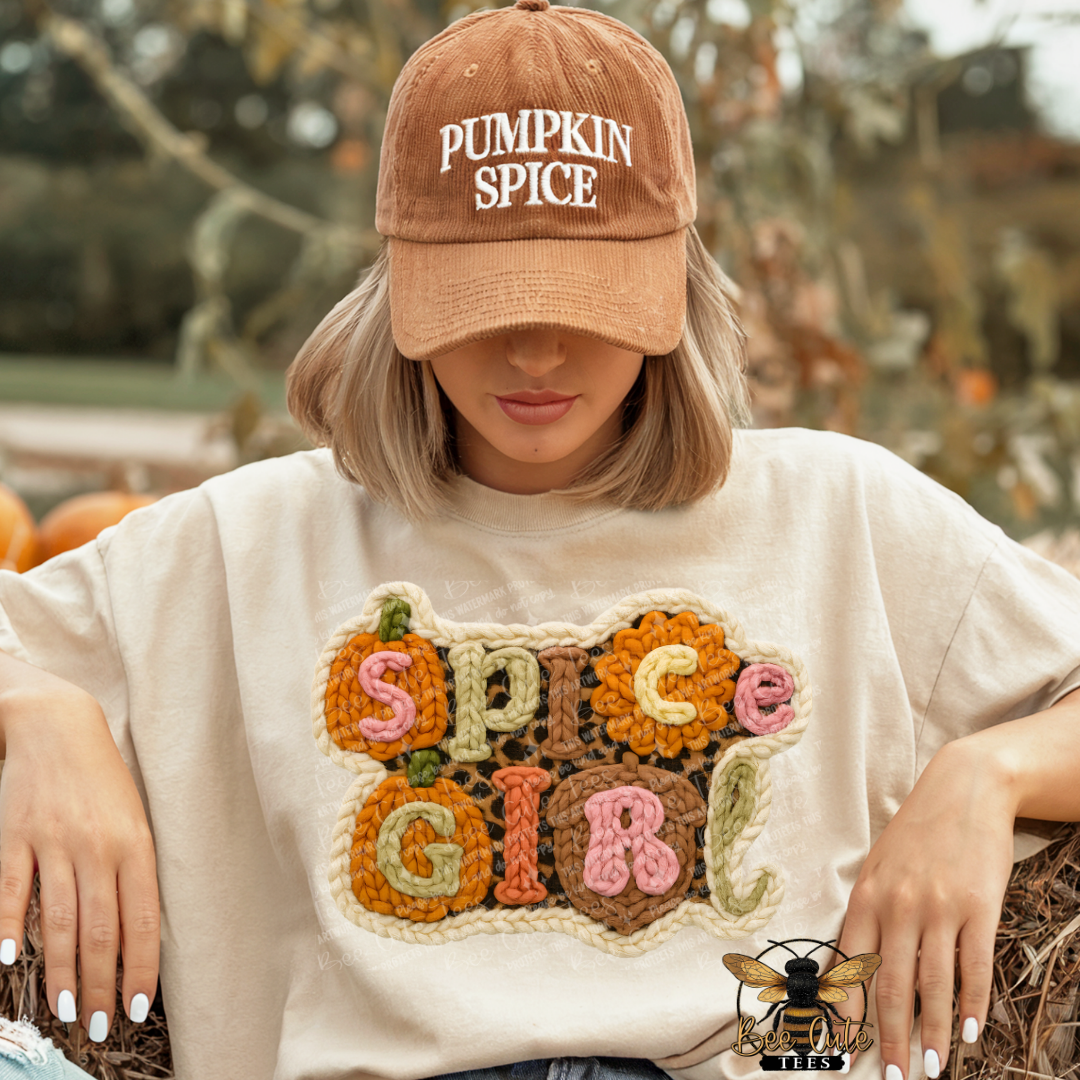 A woman wearing a tan baseball cap that says 'PUMPKIN SPICE' and a cream-colored t-shirt with a large, colorful, textured 'SPICE GIRL' graphic on the front. She is posed outdoors on a hay bale with pumpkins and a blurred autumn background.