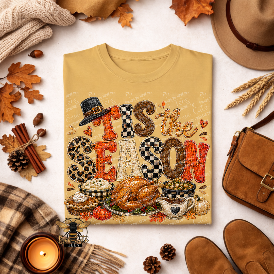 A beige graphic T-shirt laid flat on a light surface, surrounded by autumn and fall-themed props including leaves, a hat, a bag, shoes, a candle, and a scarf. The shirt reads “THIS is the SEASON” in decorative lettering with a turkey, pie, popcorn, and coffee cup illustration.