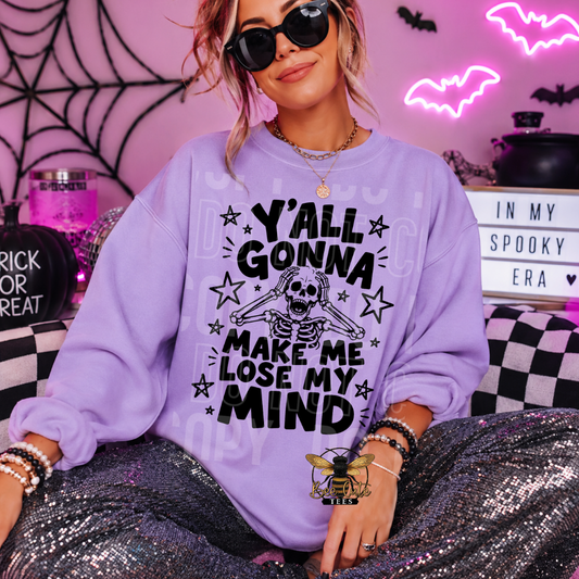 A woman wearing a lavender crewneck sweatshirt with a black skeleton and text graphic that reads 'Y'all Gonna Make Me Lose My Mind', styled for Halloween in a spooky room with neon bats and a light box.