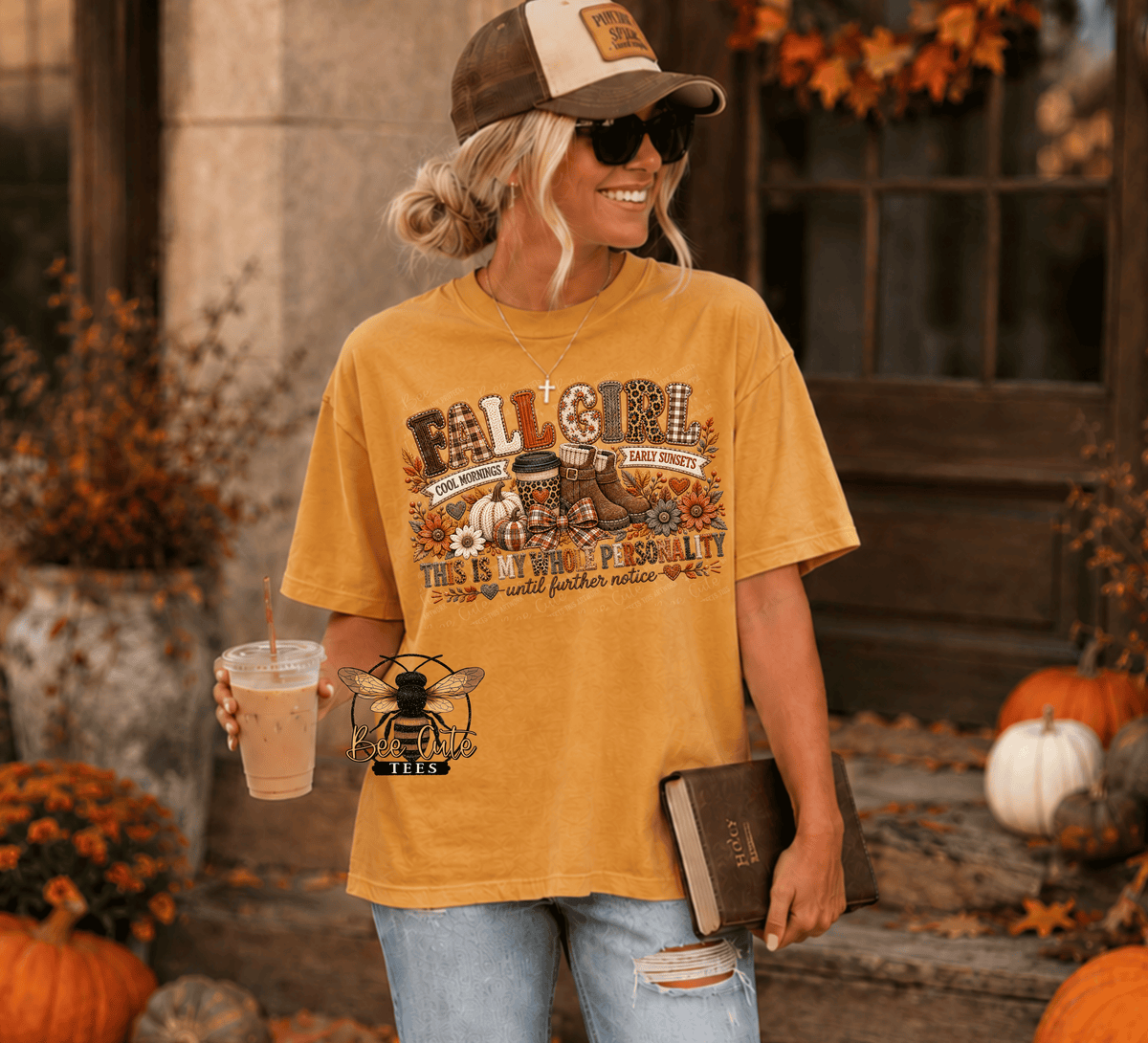 A woman wearing a mustard yellow graphic t-shirt with a fall-themed design, holding a drink and a book in an autumn-themed outdoor setting.