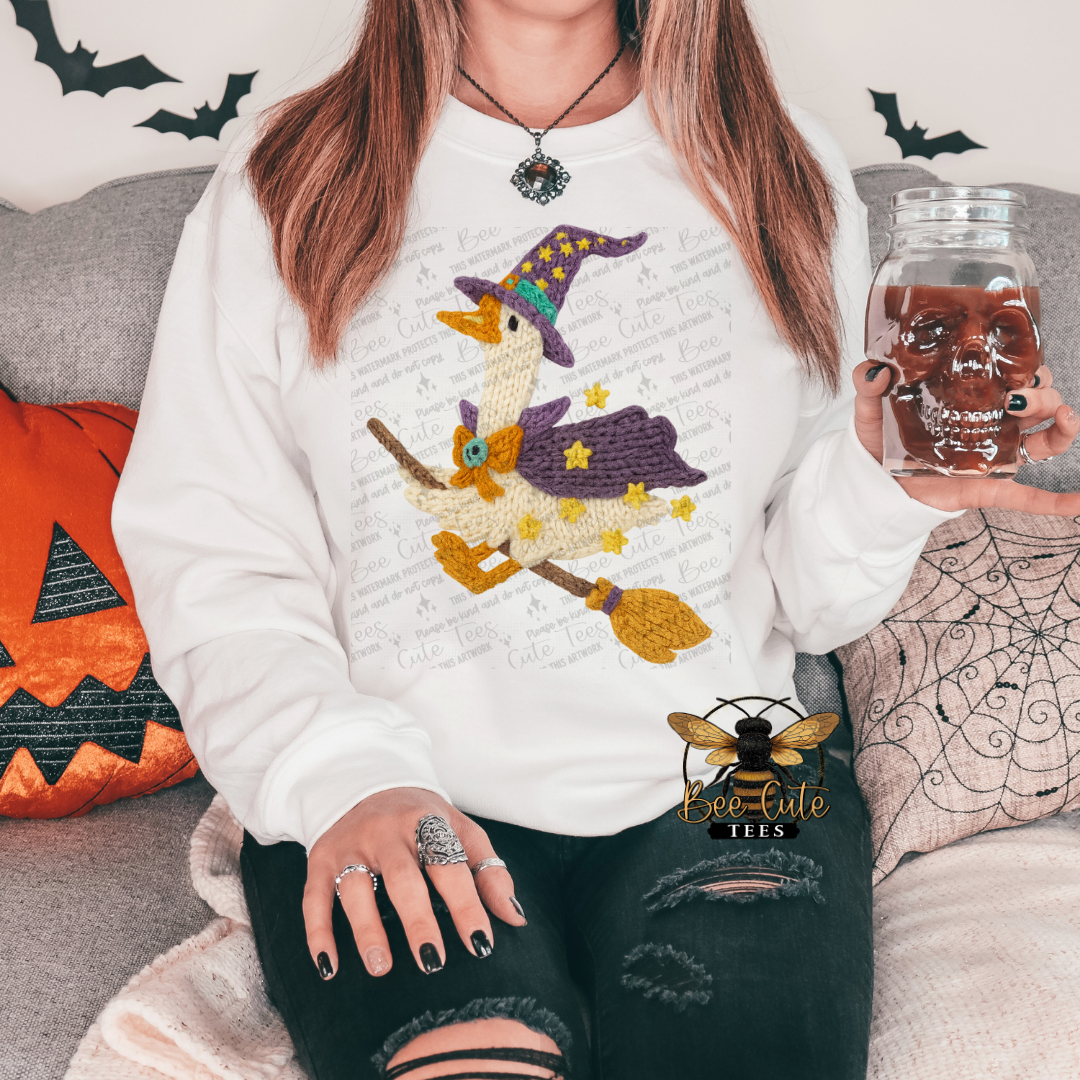 A woman wearing a white crewneck sweatshirt with a colorful embroidered witch duck graphic, sitting on a couch and holding a skull-topped mason jar.