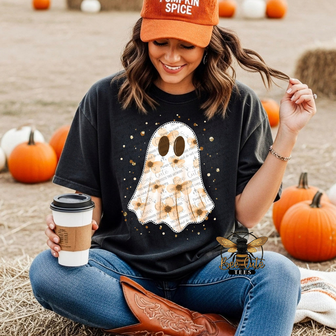 A woman sitting outdoors in a pumpkin patch wears a black graphic t-shirt with a white and brown sunflower-patterned ghost design. She is also wearing an orange baseball cap that reads 'Pumpkin Spice' and holding a coffee cup.