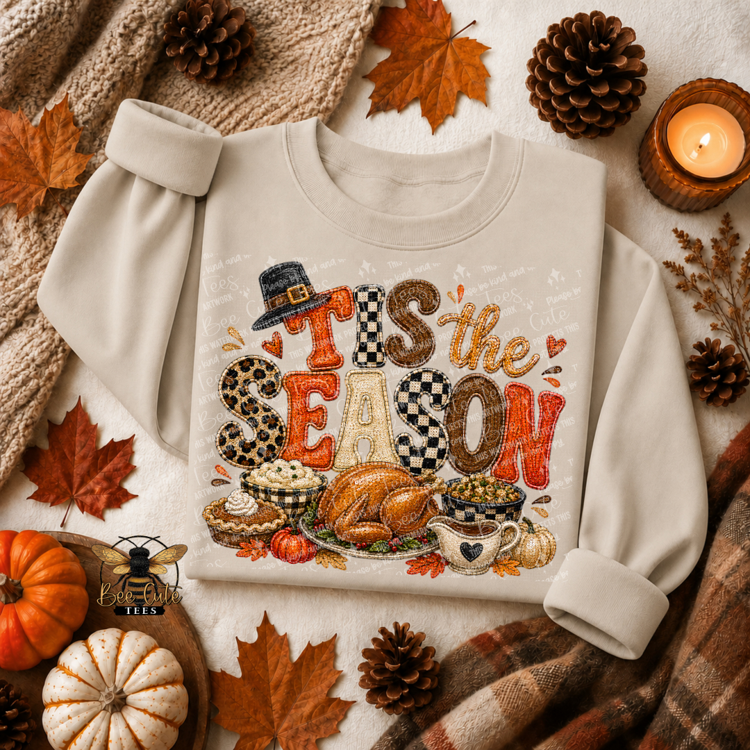 A cream-colored crewneck sweatshirt with rolled cuffs, featuring a festive autumn-themed graphic that reads 'Tis the SEASON' in patterned letters above a Thanksgiving feast illustration. The sweatshirt is styled in a flat lay with fall leaves, pinecones, a knitted blanket, a small pumpkin, and a lit candle.