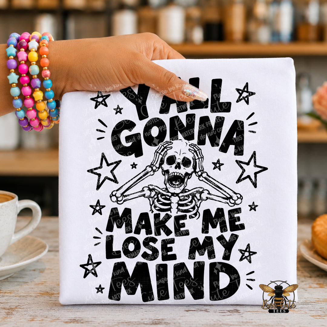 A white graphic T-shirt held by a hand wearing colorful beaded bracelets, featuring a black distressed skeleton and the text 'Y'all Gonna Make Me Lose My Mind' with star accents. The shirt is photographed in a warm, blurred kitchen or cafe setting.