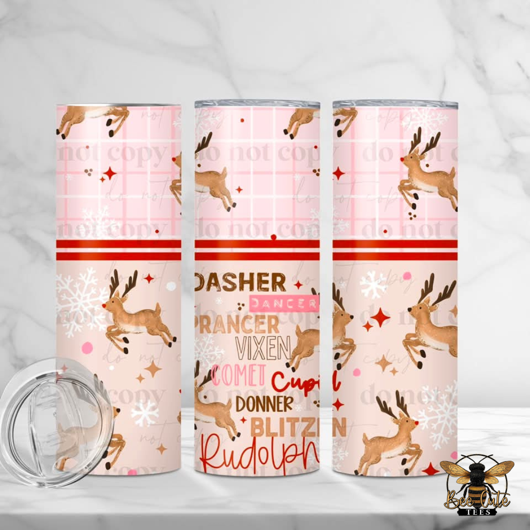 Three slim tumblers with a pink holiday-themed pattern featuring leaping deer, snowflakes, stars, and text reading 'Dasher, Prancer, Vixen, Comet, Donner, Blitz, and Rudolph' are shown on a light marble surface with a clear lid placed in front. A bee logo watermark appears in the bottom right corner.