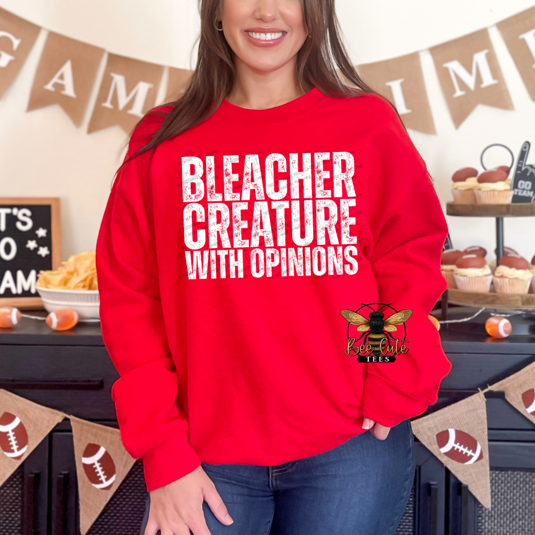 A woman wearing a bright red crewneck sweatshirt with the text 'BLEACHER CREATURE WITH OPINIONS' in distressed white letters, featuring a bee logo on the lower right. She is standing in a decorated indoor setting with football-themed bunting and cupcakes.