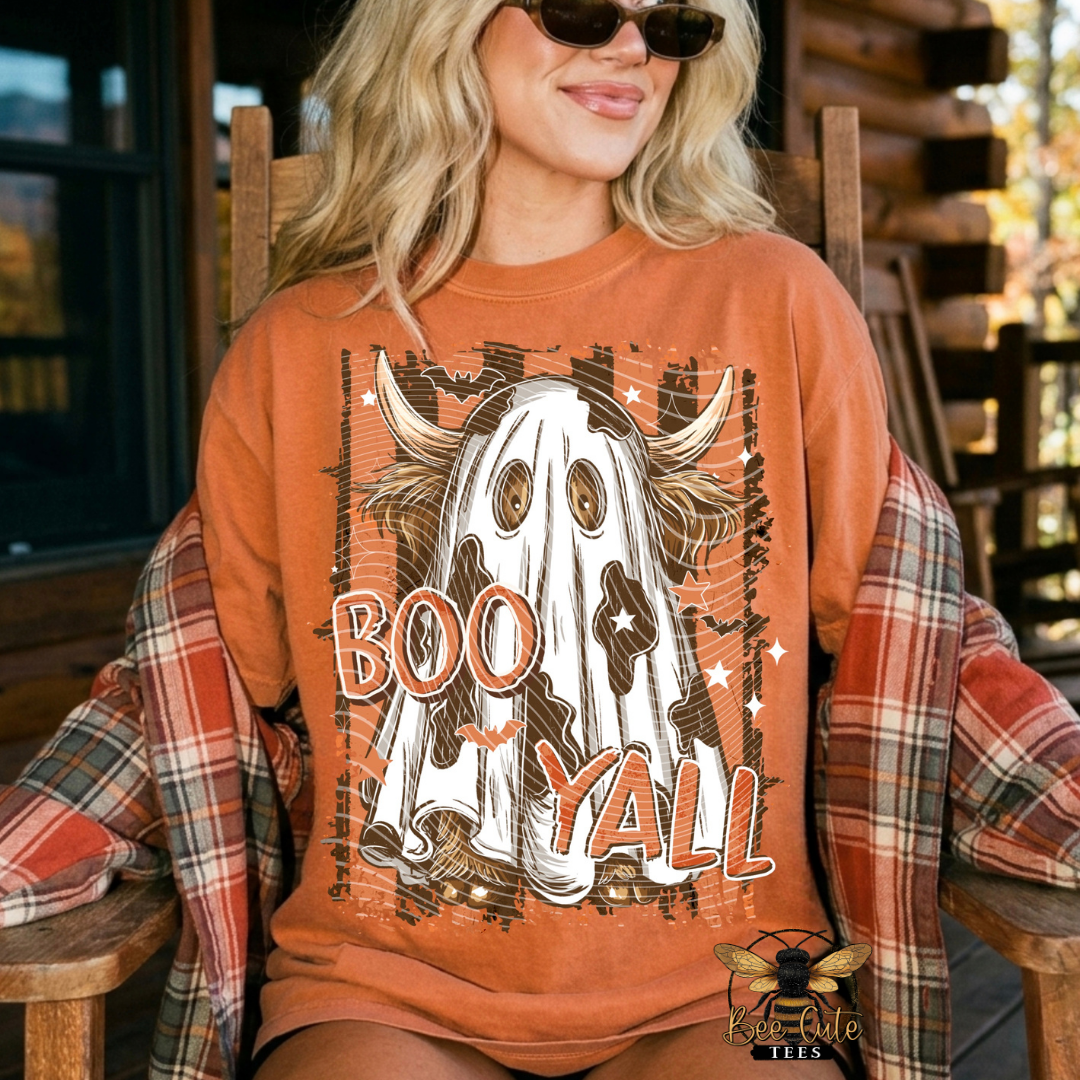 A woman wearing a rust-orange graphic sweatshirt with a ghost and the words 'BOO Y'all' sits in a wooden rocking chair. She has a red and brown plaid flannel shirt draped over her shoulders, and a 'Bee Cute Tees' logo is visible in the bottom right corner.