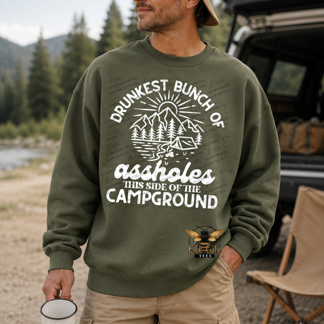 A man wearing an olive green crewneck sweatshirt with a white graphic that reads 'DRUNKEST BUNCH OF assholes THIS SIDE OF THE CAMPGROUND' and a mountain/campsite illustration. The sweatshirt also features a bee logo near the lower right front.