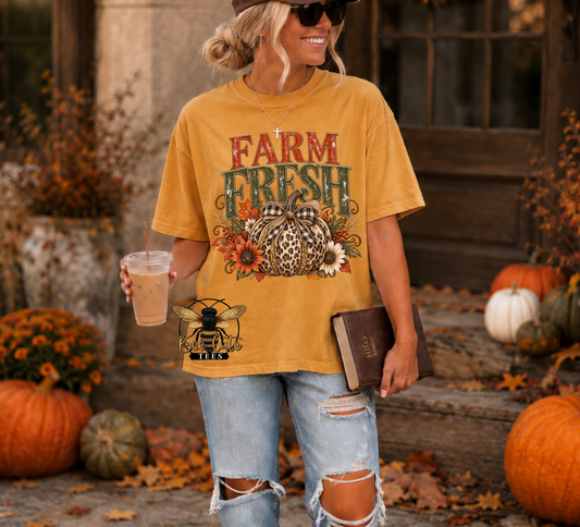 A woman wearing a mustard yellow graphic t-shirt with a 'Farm Fresh' pumpkin and floral design, paired with ripped light-wash jeans, standing in an autumn-themed outdoor setting with pumpkins and fall leaves.