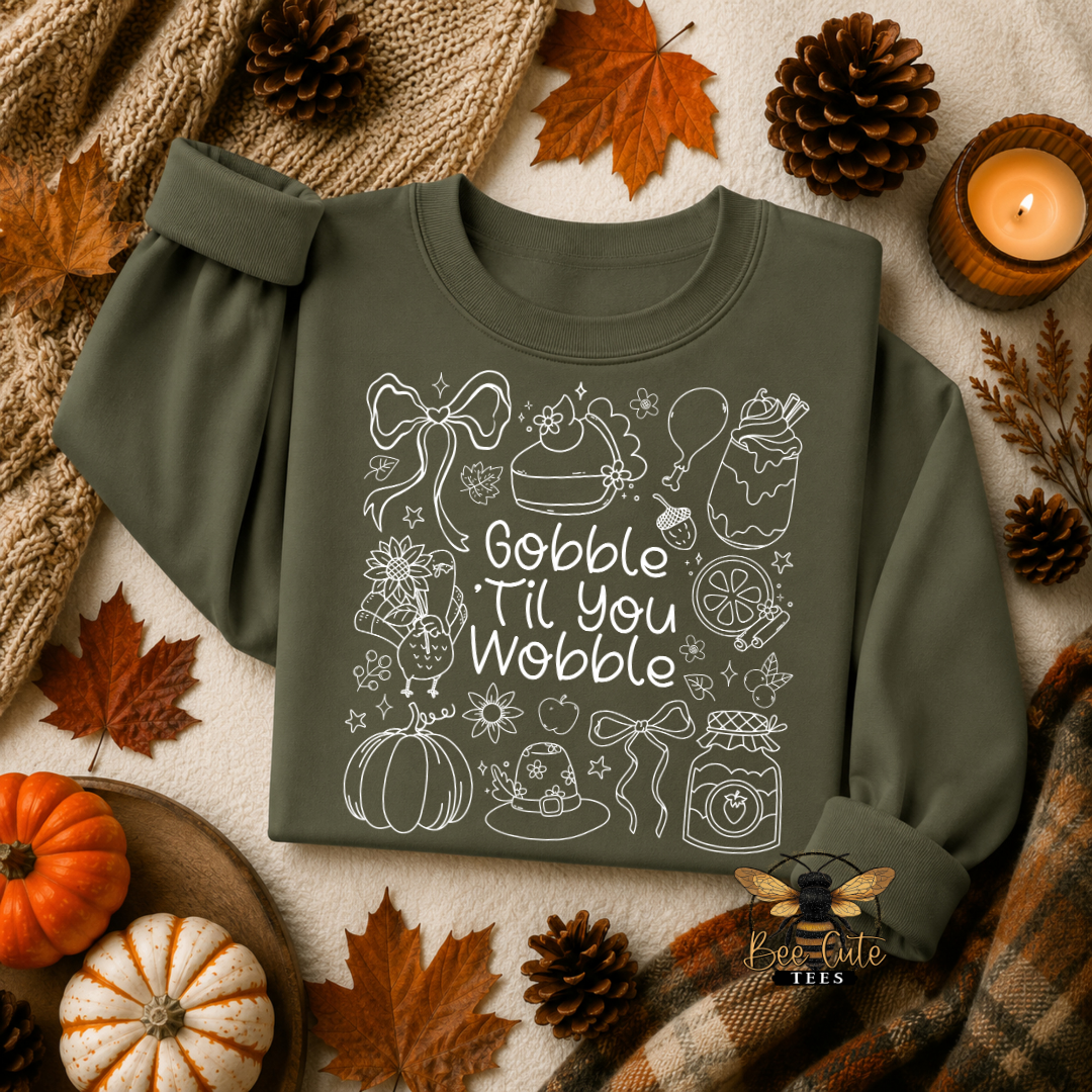 An olive green long-sleeve t-shirt with a white line-art Thanksgiving graphic and the text 'Gobble Til You Wobble' on the front, styled in a cozy autumn flat-lay with pumpkins, fall leaves, pinecones, a knitted blanket, and a lit candle.
