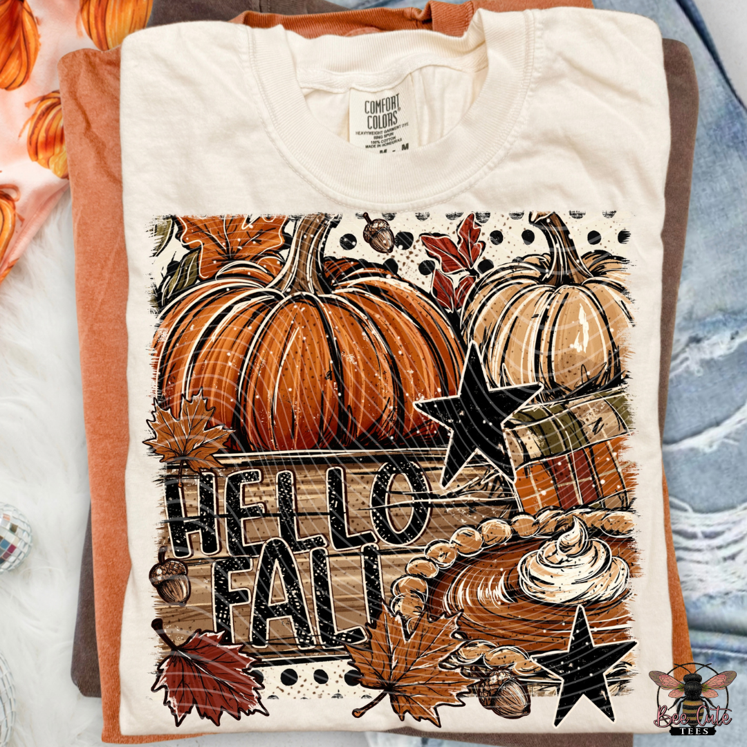 A folded cream-colored t-shirt with a detailed autumn graphic print featuring pumpkins, maple leaves, acorns, and a wooden sign that reads 'HELLO FALL'. The shirt is layered over orange and brown t-shirts on a styled flat-lay background with a blue denim shirt and orange pumpkins.