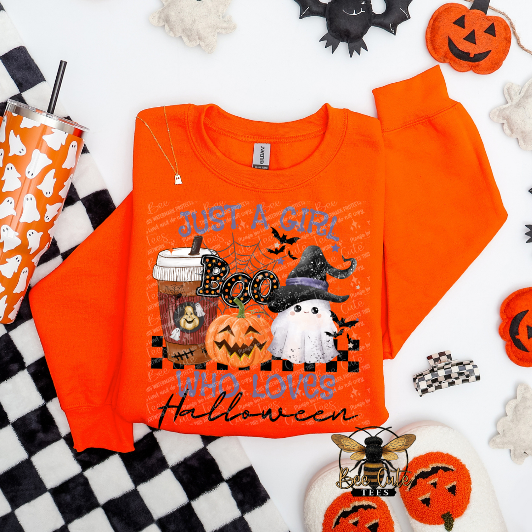 An orange crewneck sweatshirt with a Halloween-themed graphic print reading 'JUST A GIRL WHO LOVES Halloween' in text, featuring jack-o'-lanterns, a ghost, bats, and a witch hat. The sweatshirt is styled in a flat-lay arrangement with a black and white checkered blanket, a ghost-patterned tumbler, and other spooky props on a white background.