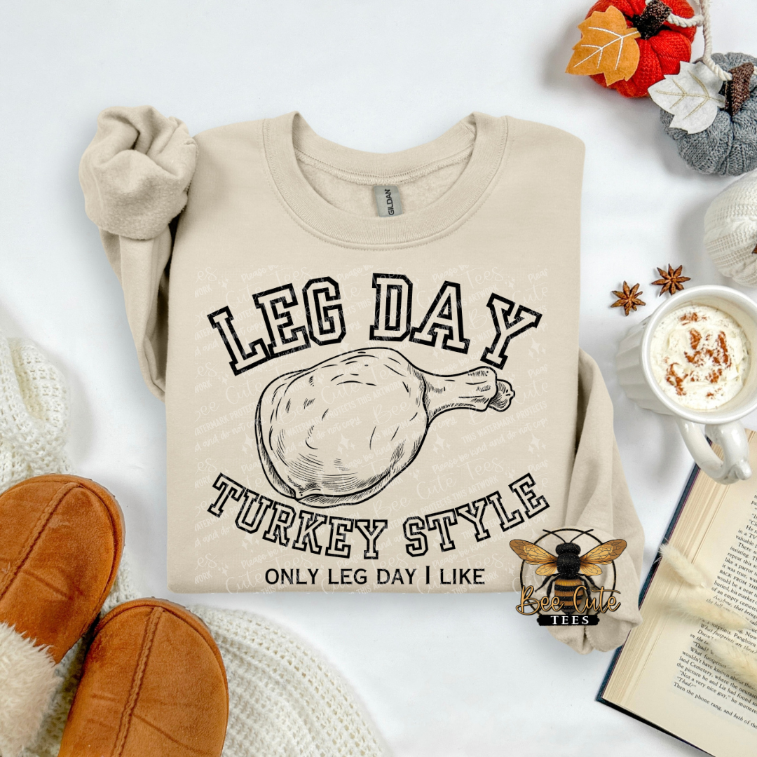A beige long-sleeve graphic sweatshirt laid flat on a white surface, featuring the text 'LEG DAY TURKEY STYLE' and 'ONLY LEG DAY I LIKE' around a turkey leg illustration. The sweatshirt is styled with cozy props including brown slippers, a mug of coffee with whipped cream, an open book, and autumn leaves.