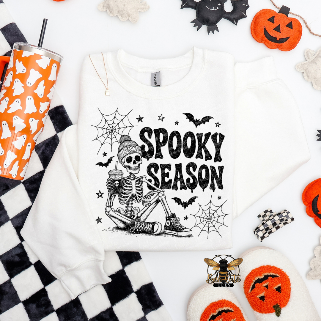 A white crewneck sweatshirt with a black 'Spooky Season' skeleton graphic, styled in a flat lay with Halloween props including a ghost-patterned tumbler, black and white checkered fabric, and orange pumpkin decorations.
