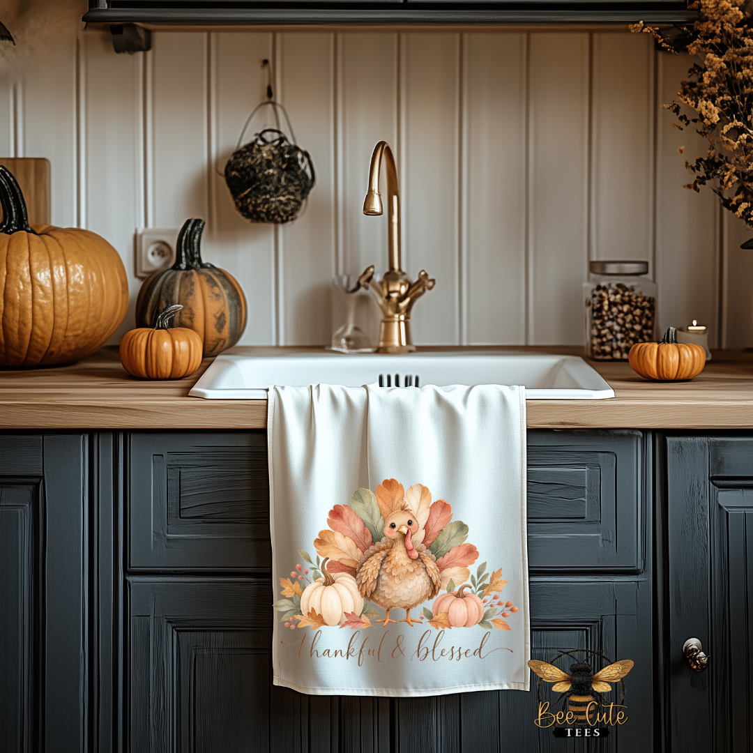 A white kitchen towel with a watercolor Thanksgiving turkey design hangs over a white sink in a rustic kitchen setting with dark cabinets, pumpkins, and a gold faucet.