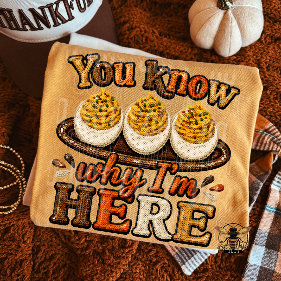 A folded yellow t-shirt, featuring a fall-themed graphic with three deviled eggs on a platter and the text 'You know why I'm HERE' in autumn colors. The item is styled on a brown knit blanket with a plaid scarf, a small pumpkin, and a beaded necklace.