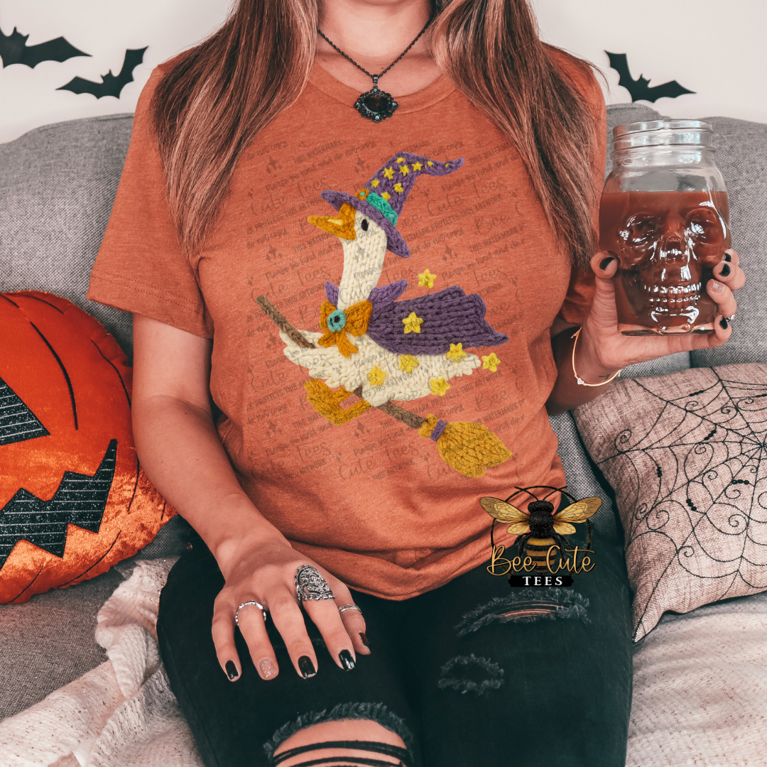 A woman sitting on a couch wearing a rust-orange t-shirt with a colorful embroidered witch duck graphic. She is holding a glass jar filled with a brown liquid, and the background shows a cozy living room setting with Halloween-themed pillows.