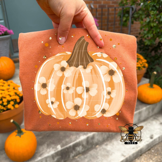 A hand holds a folded t-shirt featuring a large illustrated pumpkin with white flowers and small gold speckles on an orange background. The scene is outdoors with pumpkins, flowers, and steps visible, and a bee logo with 'Bee Cute Tees' appears in the lower right corner.