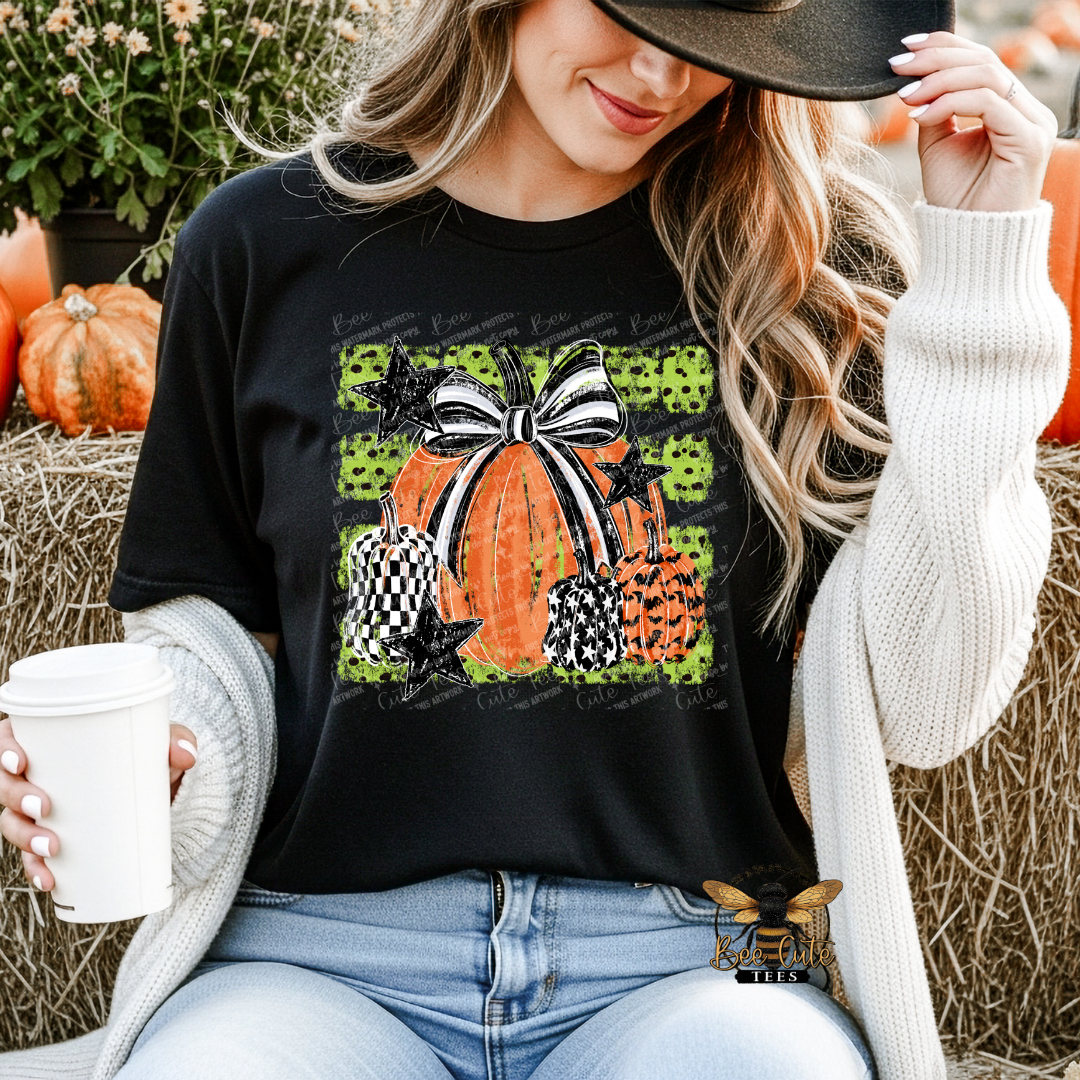 A woman wearing a black graphic t-shirt with a Halloween pumpkin design, a white knit cardigan, and a black hat, sitting in a fall-themed setting with pumpkins and hay.