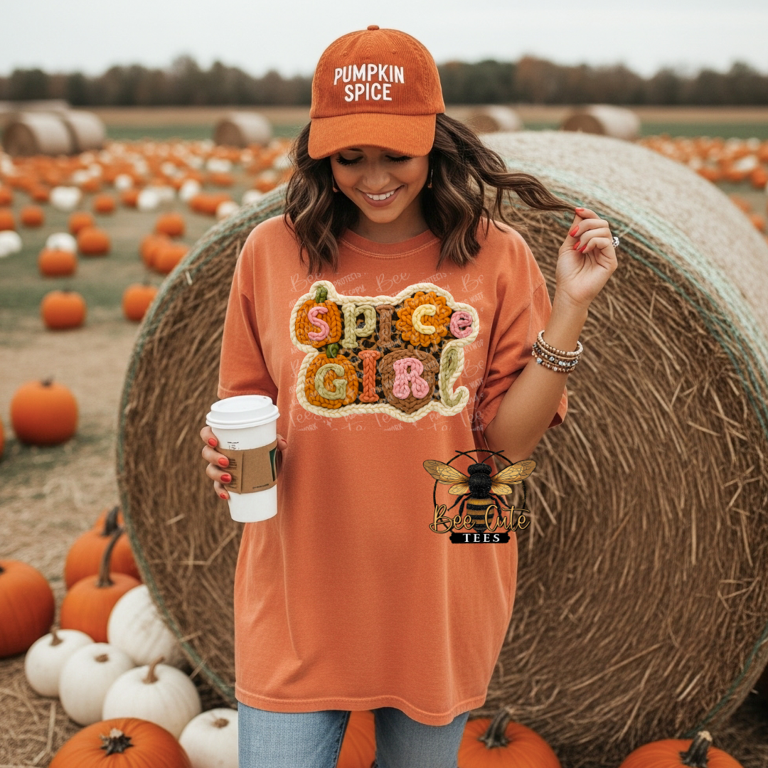 A woman standing in a pumpkin patch wearing an orange t-shirt with a 'Spice Girl' graphic made of pumpkins and a matching orange 'Pumpkin Spice' cap, holding a coffee cup next to a large hay bale.