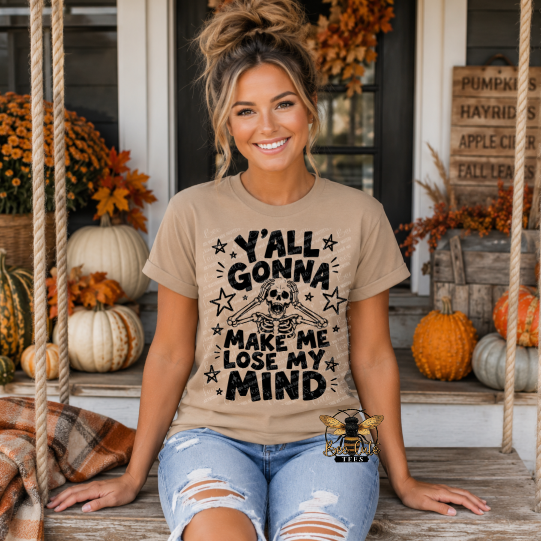 A woman sitting on a porch swing wearing a beige graphic t-shirt with a black skull and stars design that reads 'Y'ALL GONNA MAKE ME LOSE MY MIND'.