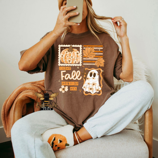 A woman sitting in a chair takes a mirror selfie while wearing a brown graphic t-shirt with a fall-themed design featuring pumpkins, leaves, and a ghost, paired with light gray sweatpants and white socks.