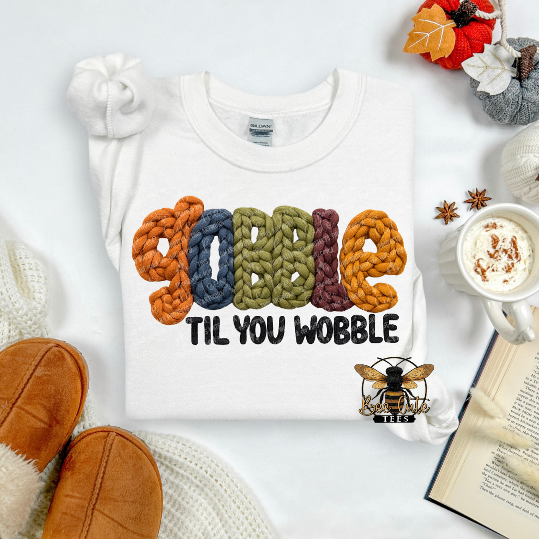 A white crewneck sweatshirt laid flat on a white surface, featuring a colorful graphic that reads 'GOBBLE' in knitted-style letters above 'TIL YOU WOBBLE' in black. The sweatshirt is styled with autumn props including orange slippers, a mug of coffee, an open book, and a small bee graphic.