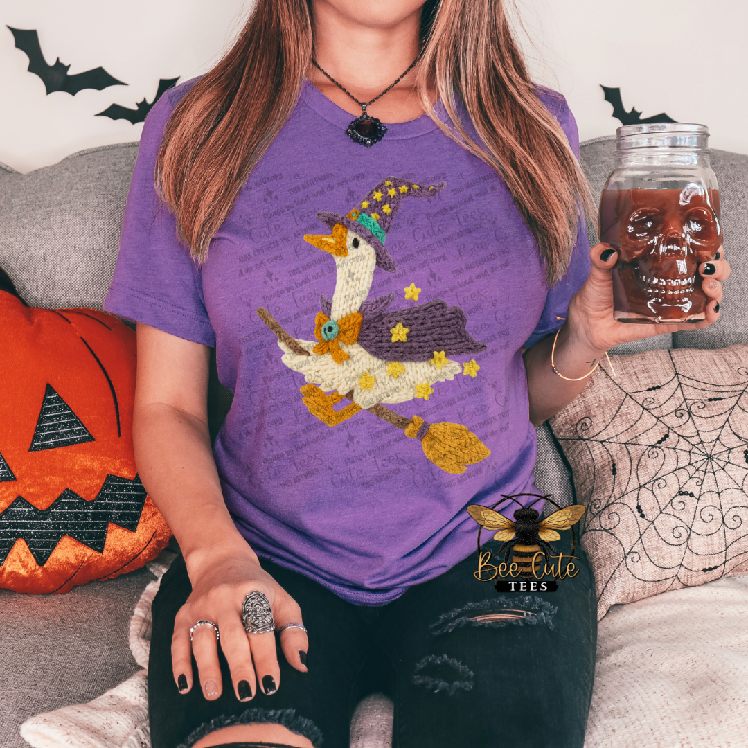A woman wearing a purple graphic t-shirt with a witch riding a broomstick design, holding a mason jar with a skull lid. She is sitting on a couch with Halloween-themed pillows.
