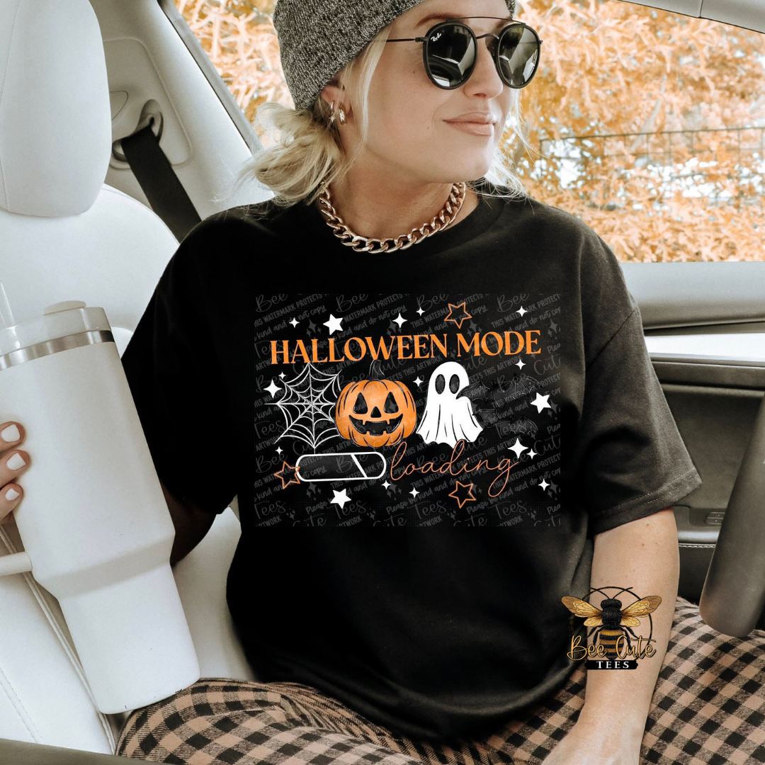 A woman sitting in a car wearing a black graphic t-shirt with a Halloween-themed design that reads 'HALLOWEEN MODE' and shows a pumpkin, ghost, spiderwebs, and a battery icon. She is also wearing a gray beanie, sunglasses, and a gold chain necklace.