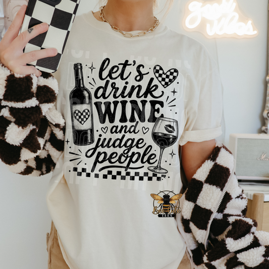 A person wearing a white graphic t-shirt with a black and white design that reads 'let's drink WINE and judge people' with wine bottle and glass illustrations. The person is holding a checkered phone case and wearing fuzzy black-and-white cardigan sleeves in a brightly lit indoor setting.