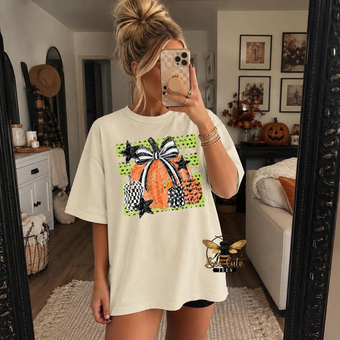 A woman takes a mirror selfie wearing an oversized cream-colored t-shirt with a colorful Halloween pumpkin graphic on the front. The graphic features orange pumpkins, black and white checkered patterns, and a black and white bow, set against a bright green polka-dot background.