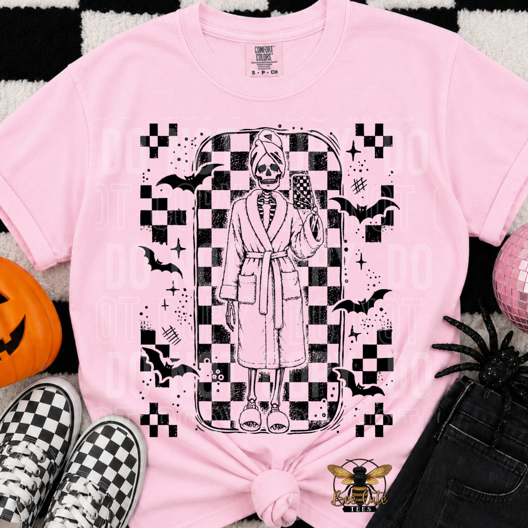 A pink graphic T-shirt laid flat with a black-and-white skeleton figure in a robe holding a phone, surrounded by bats, checkered motifs, and spooky details. The shirt is styled with black-and-white sneakers, a jack-o'-lantern, a spider, jeans, and a pink disco ball on a black-and-white patterned surface.
