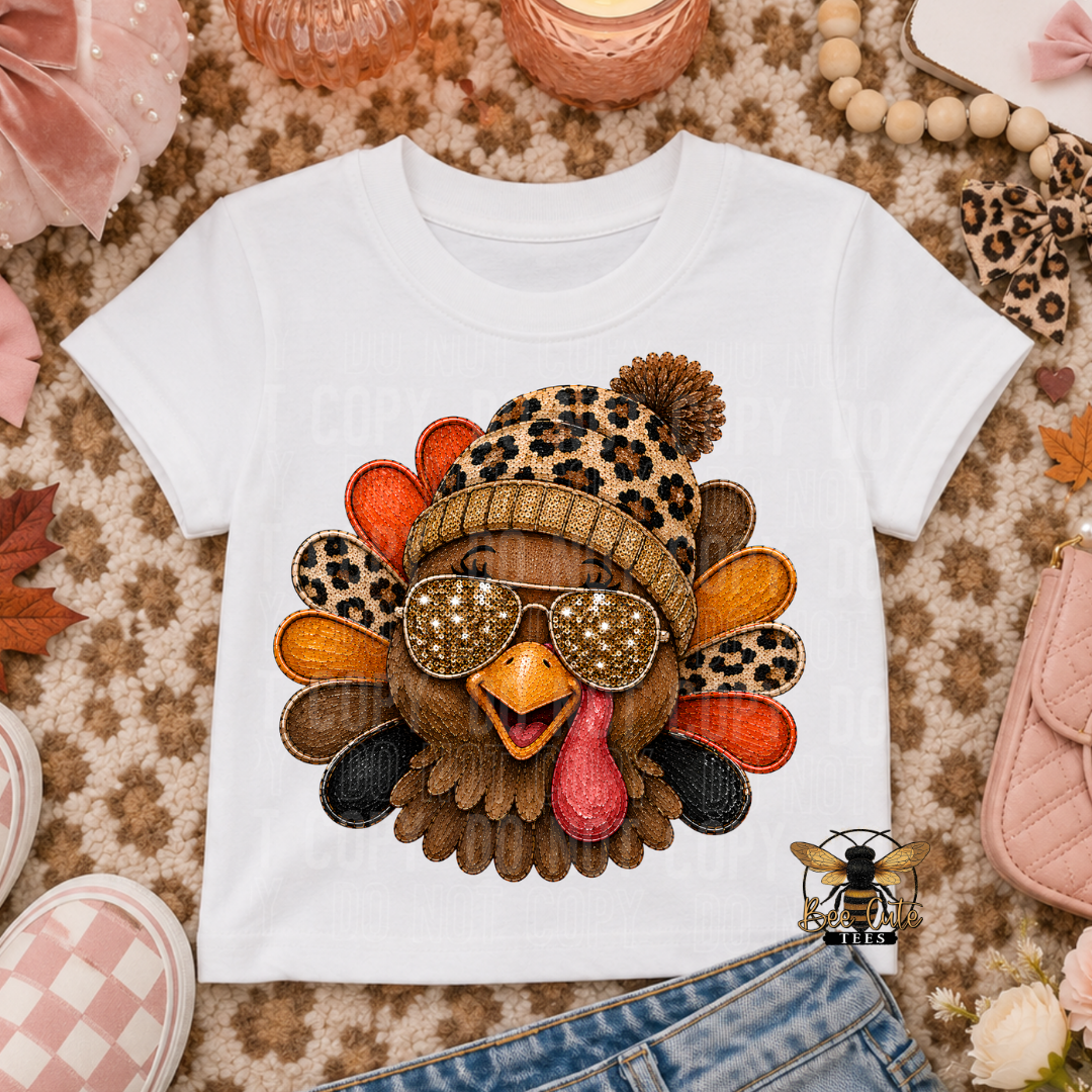A white short-sleeve T-shirt laid flat on a patterned rug, featuring a large turkey graphic with a leopard-print beanie, starry sunglasses, and orange and brown feathers. The shirt is styled with jeans, pink shoes, pumpkins, beads, and autumn leaves around it.
