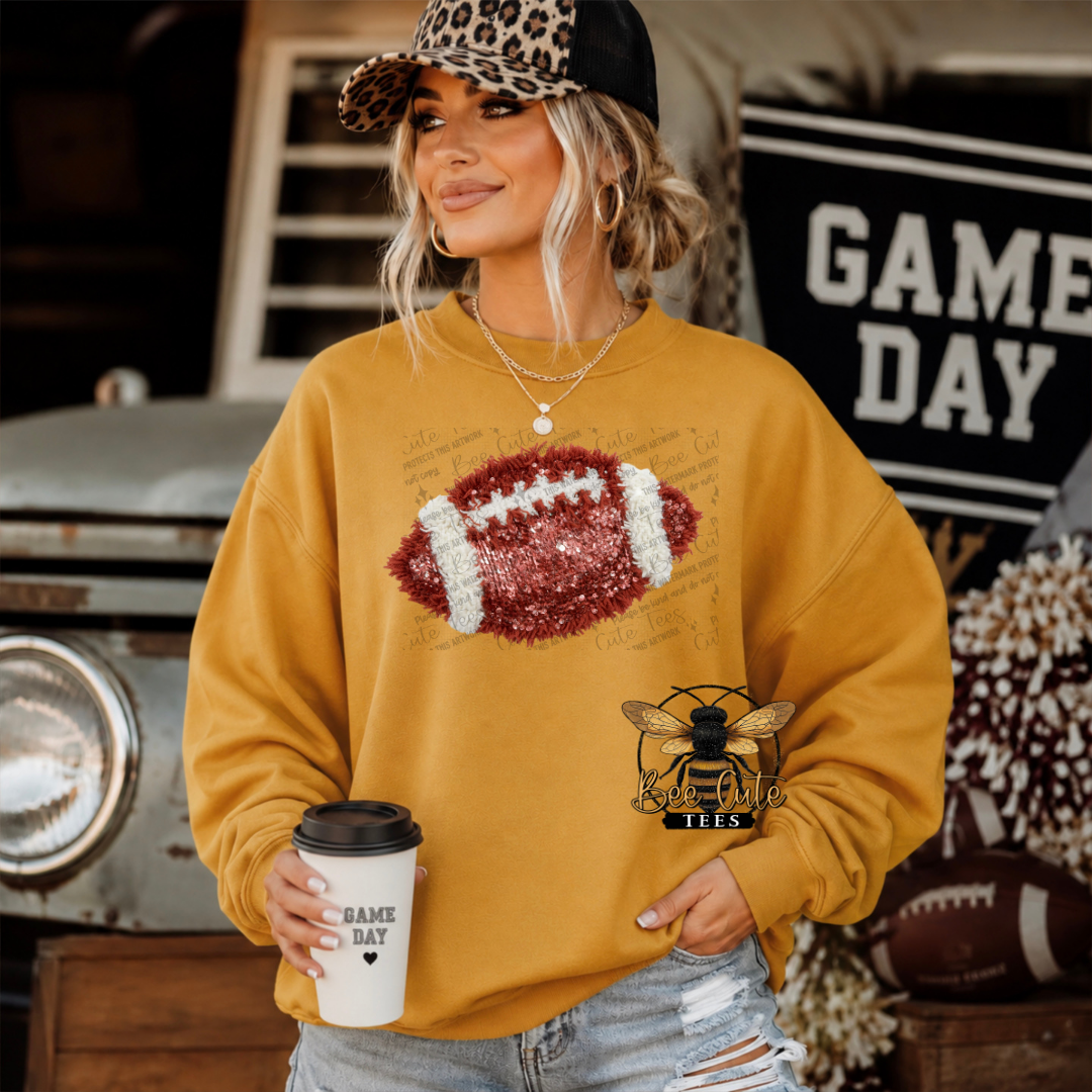 A woman wearing a mustard yellow sweatshirt with a football graphic on the chest and a bee logo on the front pocket, holding a coffee cup in a rustic outdoor setting.