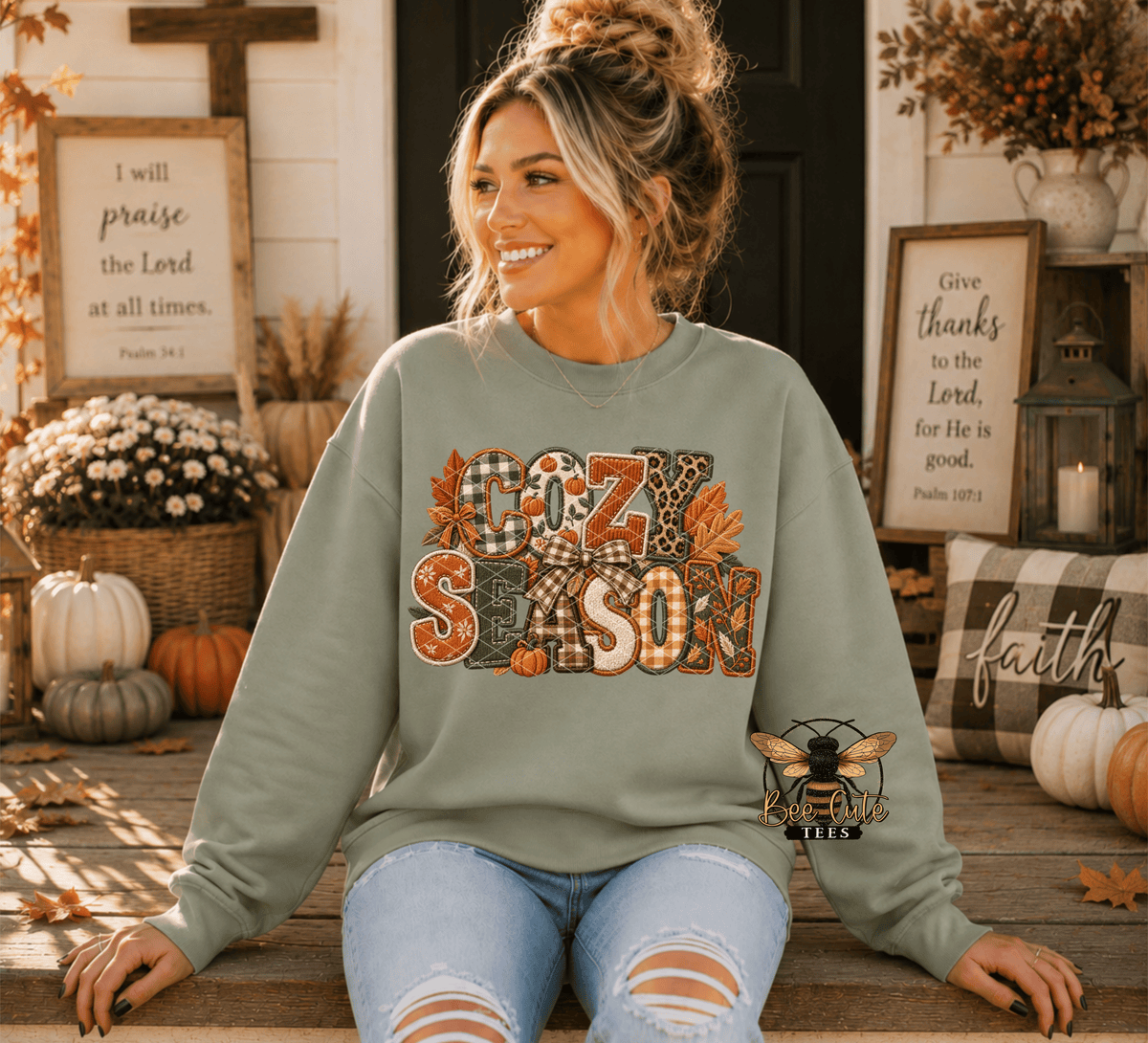 A woman sitting on a wooden porch wearing a sage green crewneck sweatshirt with a 'COZY Season' autumn graphic and a bee patch. The background features fall decorations including pumpkins, a cross, and framed scripture.