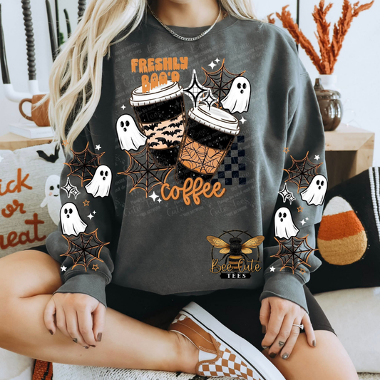 A woman wearing a dark gray crewneck sweatshirt with a Halloween-themed coffee graphic. The design features two iced coffee cups, white ghosts, spiderwebs, and the text 'FRESHLY BAKED coffee'.