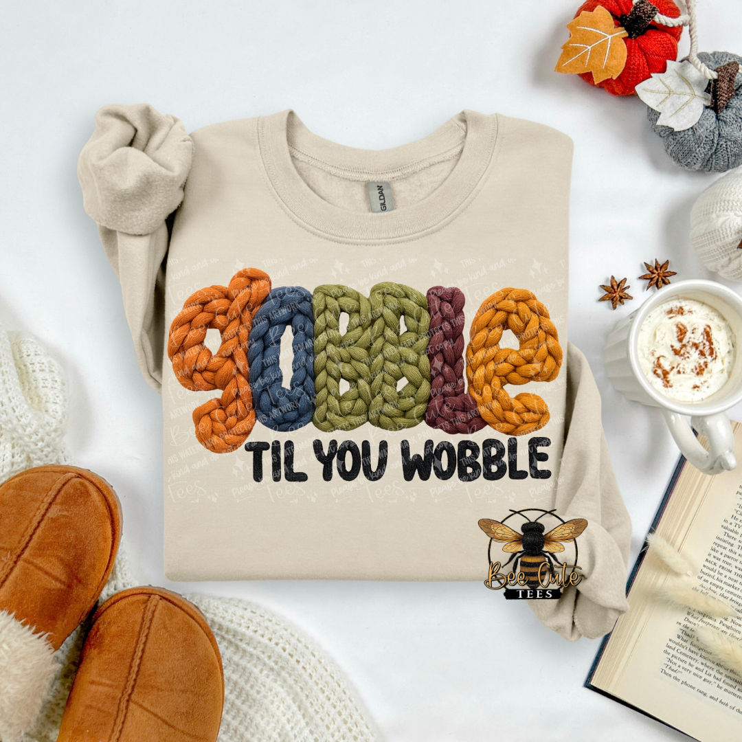A beige crewneck sweatshirt laid flat on a white surface, featuring the phrase 'YOBBLE TIL YOU WOBBLE' in colorful chunky-knit lettering. The layout is styled with cozy autumn props including a cup of coffee with whipped cream, an open book, brown slippers, and a small bee logo with the text 'BEE CUTE TEES'.