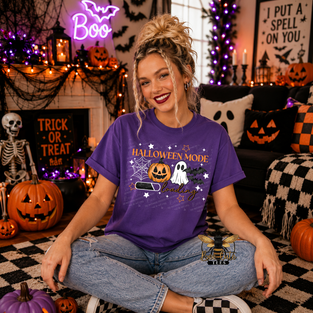 A woman sits cross-legged in a decorated Halloween living room wearing a purple graphic T-shirt that reads 'HALLOWEEN MODE' with a jack-o'-lantern, ghost, bat, and stars. The room has pumpkins, a skeleton decor, a neon 'Boo' sign, and black-and-white checkered flooring.