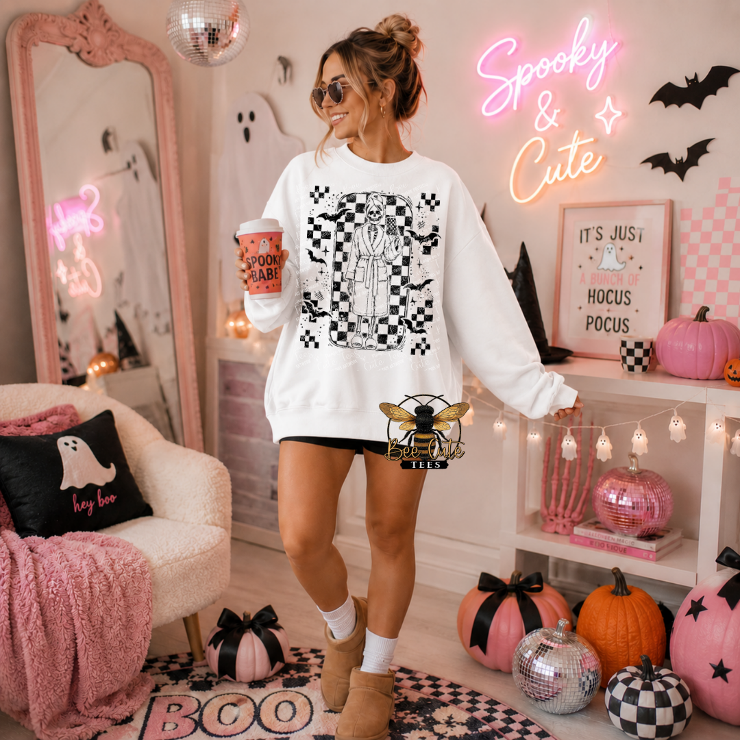A woman stands in a decorated Halloween-themed room wearing a white crewneck sweatshirt with a black and white skeleton graphic. She holds a coffee cup and wears black shorts, white socks, and tan boots.