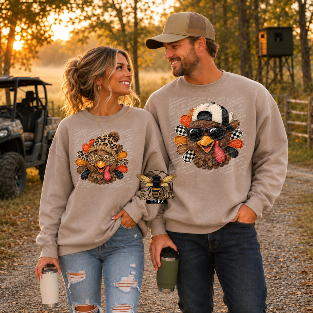 A man and a woman standing outdoors in a wooded setting at sunset, both wearing matching beige crewneck sweatshirts with a large turkey graphic on the front. The woman's sweatshirt has a smaller turkey graphic, and the man's has a larger turkey wearing sunglasses and a baseball cap.