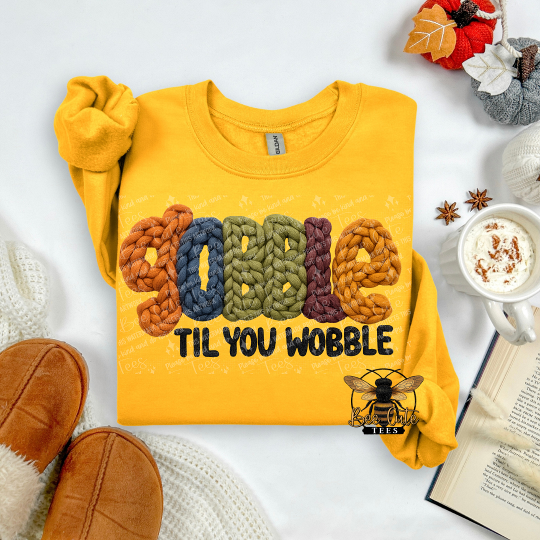 A folded yellow sweatshirt laid flat on a white surface with autumn-themed props. The sweatshirt has a colorful knitted-style 'GOBBLE' graphic and the phrase 'TIL YOU WOBBLE' in black.