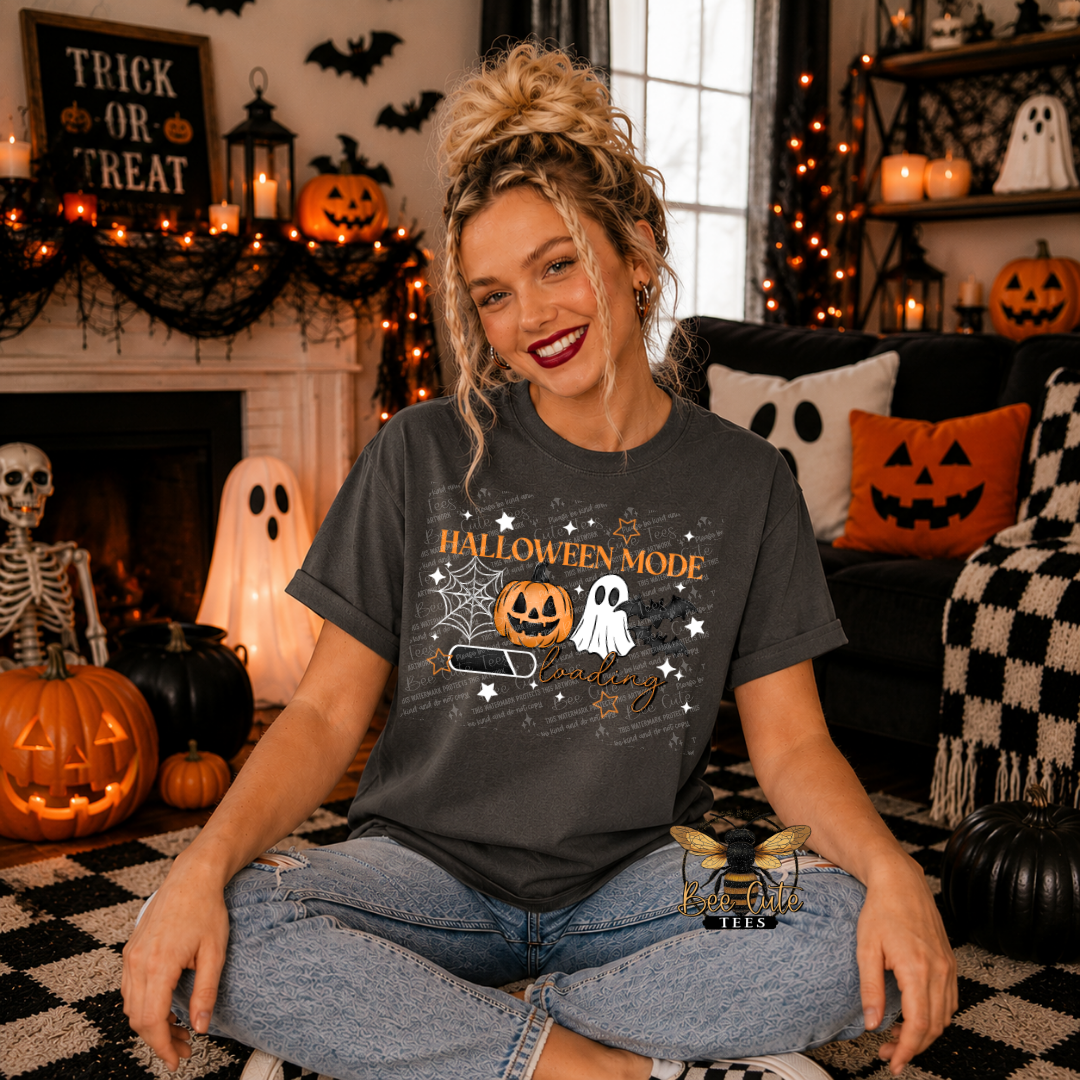 A woman sitting cross-legged in a cozy, decorated Halloween living room, wearing a dark gray graphic t-shirt that reads 'HALLOWEEN MODE' with a jack-o'-lantern, ghost, and bat design.