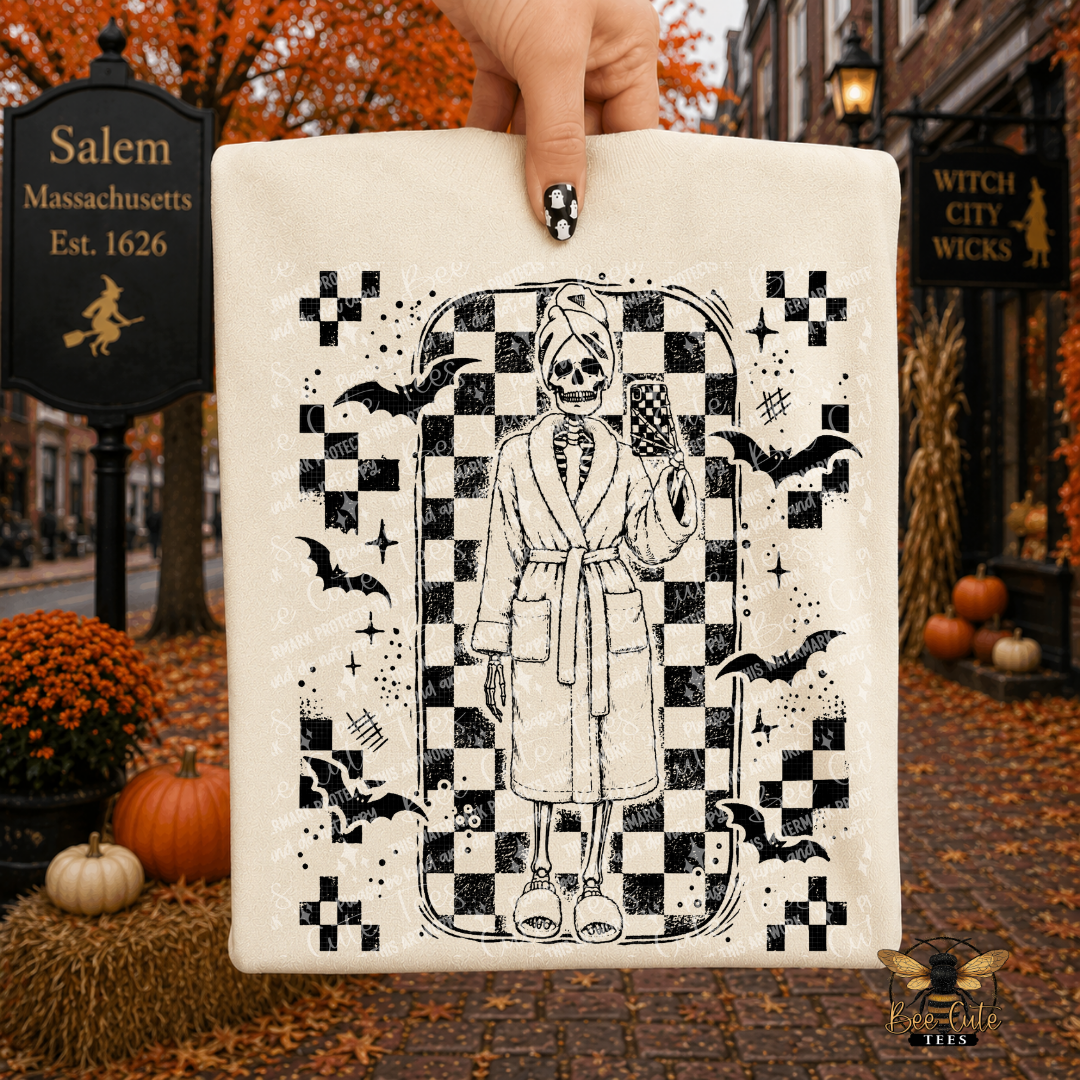 A hand holds a cream-colored square canvas print featuring a black-and-white illustration of a skeleton in a robe taking a mirror selfie, surrounded by bats and checkered patterns. The background is a Halloween-themed street scene in Salem, Massachusetts, with autumn leaves, pumpkins, and storefronts.
