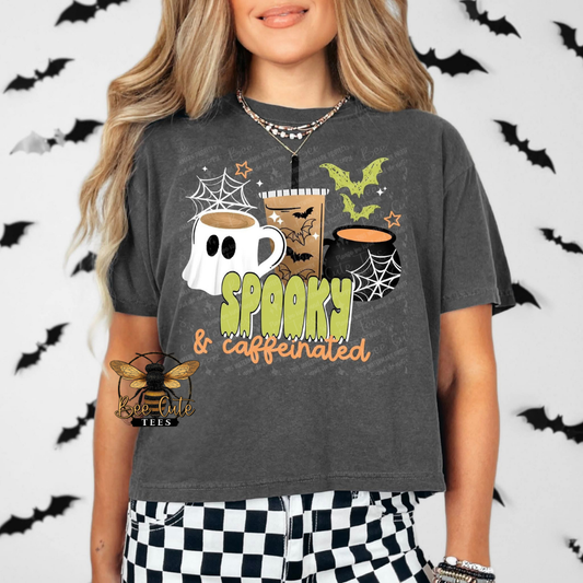 A woman wearing a dark gray graphic t-shirt with a 'Spooky & Caffeinated' Halloween design featuring coffee cups, a ghost, spiderwebs, and bats. The background is white with black bat silhouettes.