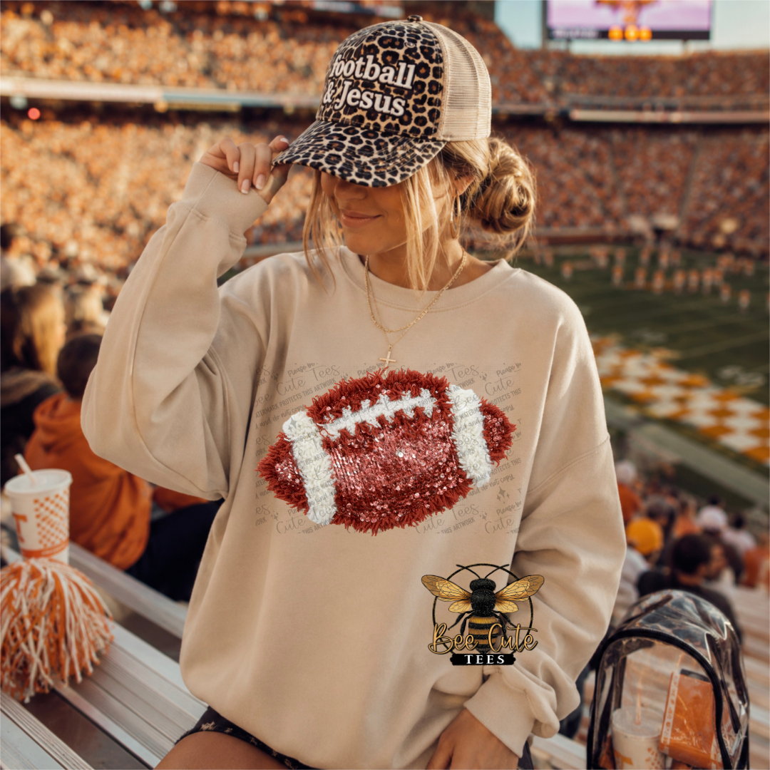 A woman in a stadium wearing a beige sweatshirt with a sequined football graphic, a leopard-print trucker hat that says 'Football Jesus,' and a bee logo on the sleeve. The background shows a crowded football field and stands.
