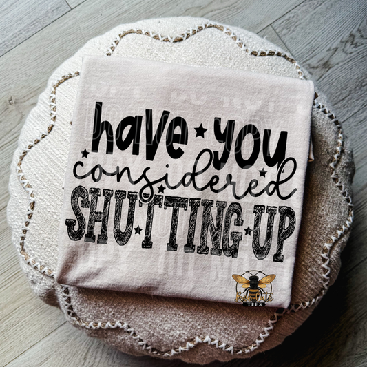 A folded light beige t-shirt with black text reading 'have you considered SHUTTING UP' and a small bee logo, placed on top of a round woven cushion over a wood floor.