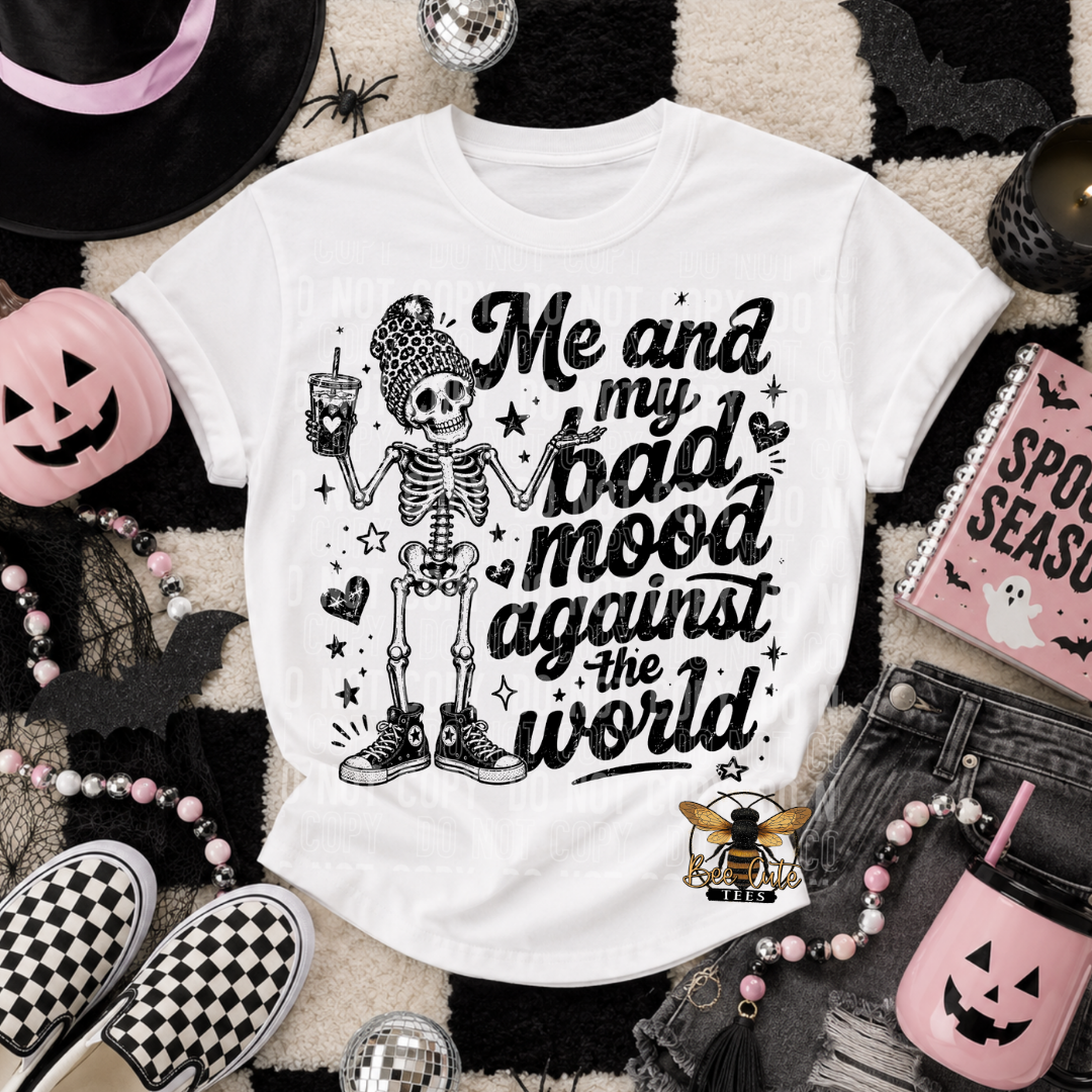 A white graphic t-shirt featuring a black and white skeleton holding a drink next to the text 'Me and my bad mood against the world'. The shirt is styled in a flat-lay arrangement with Halloween-themed props including a jack-o'-lantern cup, checkered sneakers, a pink pumpkin, and a beaded necklace on a black and white patterned rug.