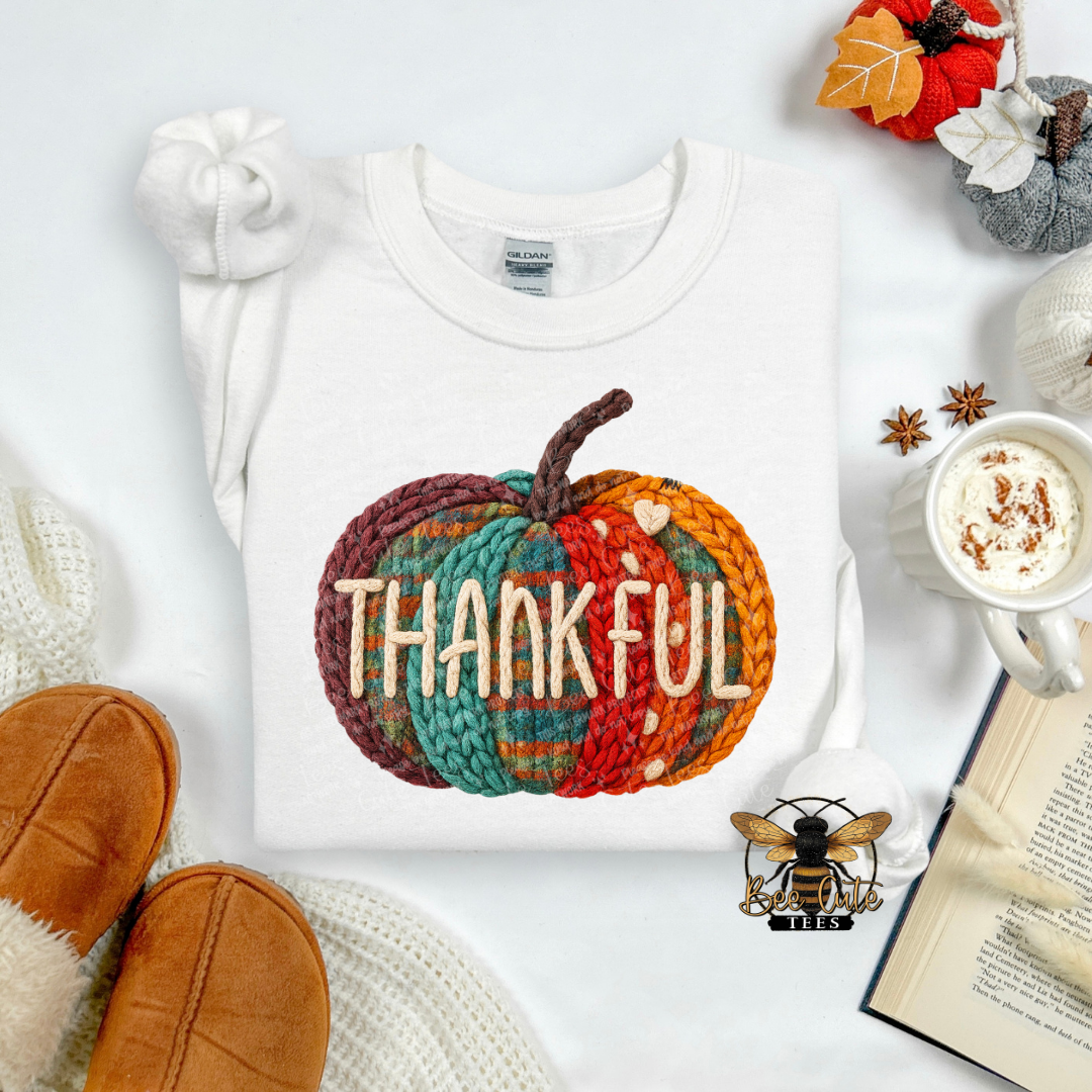 A folded white crewneck sweatshirt with a colorful autumn-themed knit pumpkin graphic and the word 'THANKFUL' across the front, styled in a cozy flat lay with brown slippers, a mug of coffee, an open book, and fall leaves.