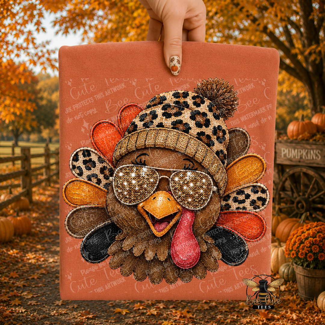 A hand holds a folded orange t-shirt featuring a stylized turkey face with a leopard-print beanie, glittery sunglasses, and a feather pattern on an orange background, set against an autumn outdoor backdrop with pumpkins and fall foliage.