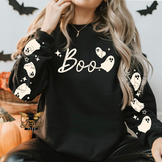 A woman wearing a black long-sleeve sweatshirt with a white 'Boo' script and ghost graphics on the sleeves and chest, posed in a Halloween-themed setting with pumpkins and bat cutouts.